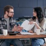 Un bărbat și o femeie stau de vorbă la masa din bucătărie, cu hârtii și căni de cafea în față