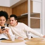 Un bărbat și o femeie stau la masa din bucătărie și se uită împreună la un telefon