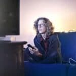 Tânăr pe canapea, cu telefonul în mână, în fața televizorului, seara