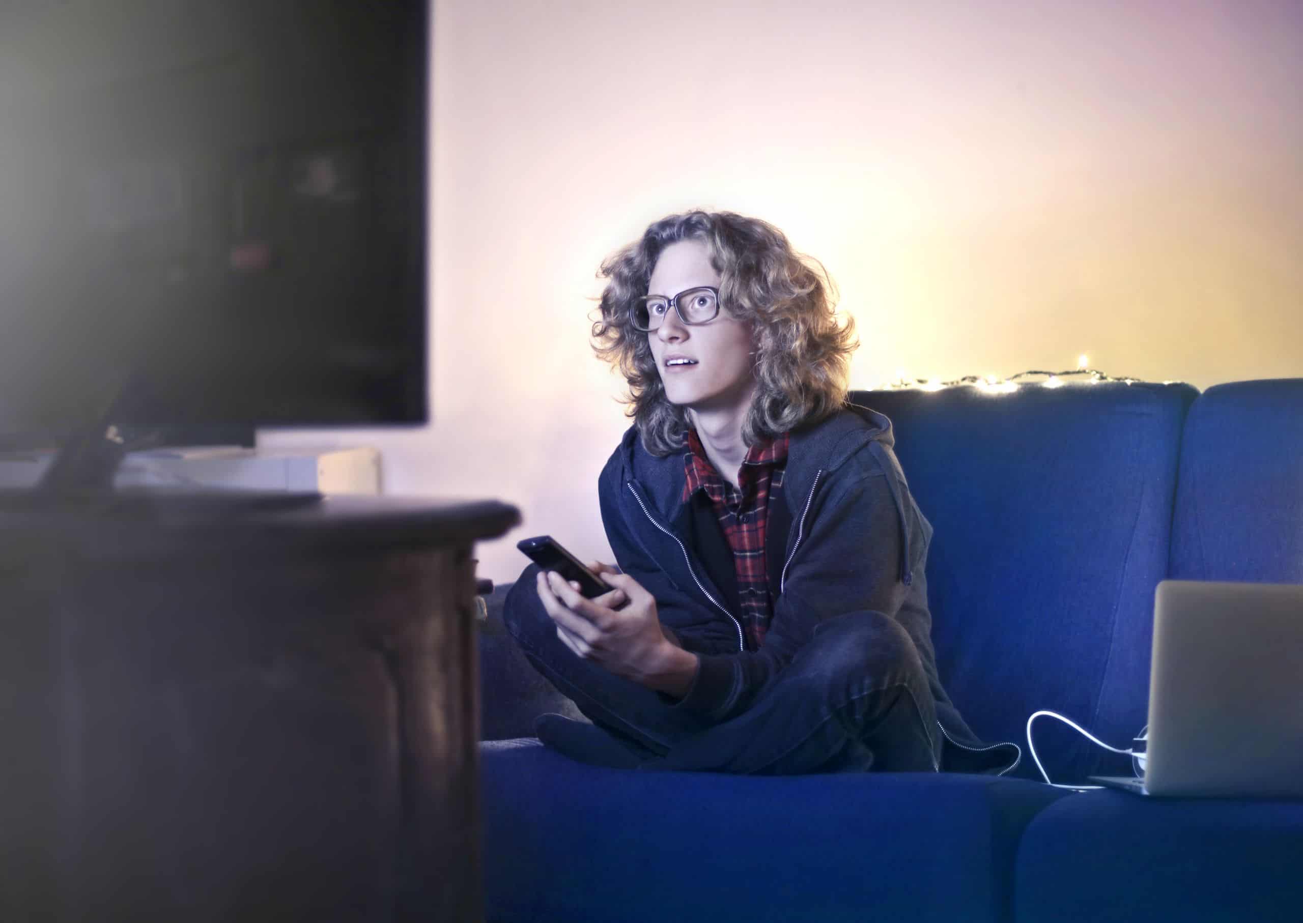 Tânăr pe canapea, cu telefonul în mână, în fața televizorului, seara