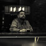 Bărbat așezat singur într-un bar întunecat, cu un pahar în mână, pierdut în gânduri