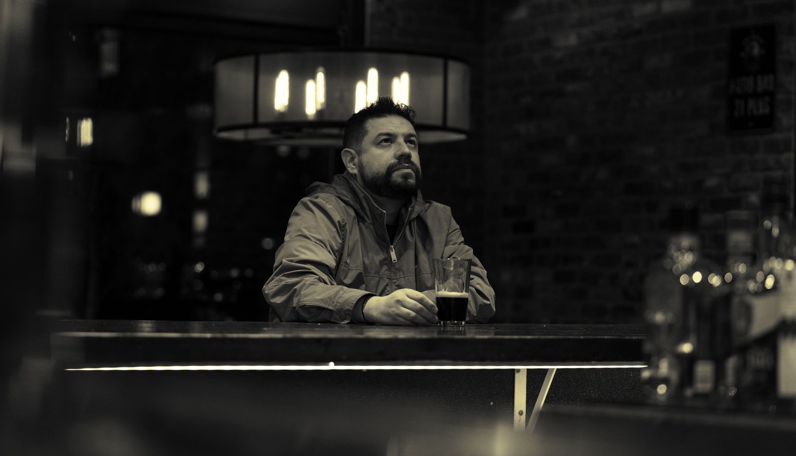 Bărbat așezat singur într-un bar întunecat, cu un pahar în mână, pierdut în gânduri