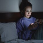 Femeie așezată pe pat, noaptea, cu fața luminată de ecranul telefonului