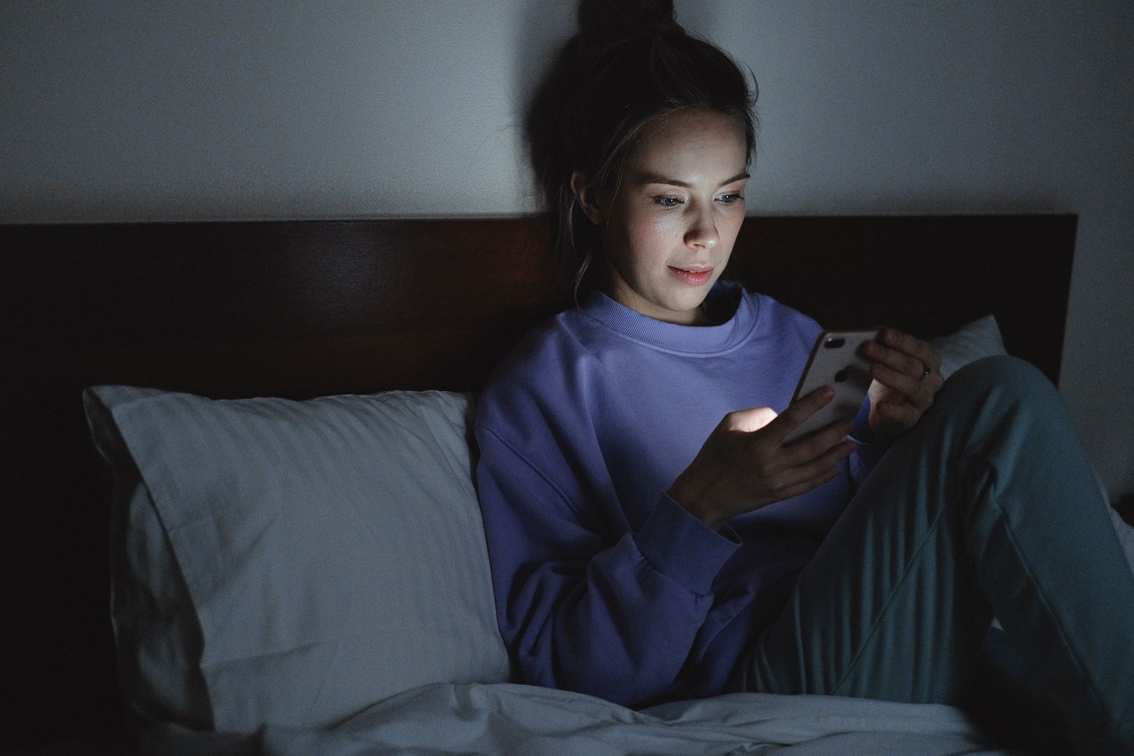 Femeie așezată pe pat, noaptea, cu fața luminată de ecranul telefonului