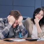 Bărbat cu capul în mâini la masa din bucătărie, cu telefon și hârtii, lângă o femeie care îl privește îngrijorată