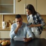 Bărbat care citește îngrijorat o scrisoare la masa din bucătărie, cu partenera aplecată lângă el
