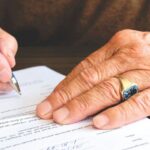 Mâna unei persoane în vârstă, cu un inel de aur, semnează un document cu pixul