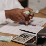 Mâini care numără bancnote pe un birou de lemn, lângă un calculator, acte și un laptop