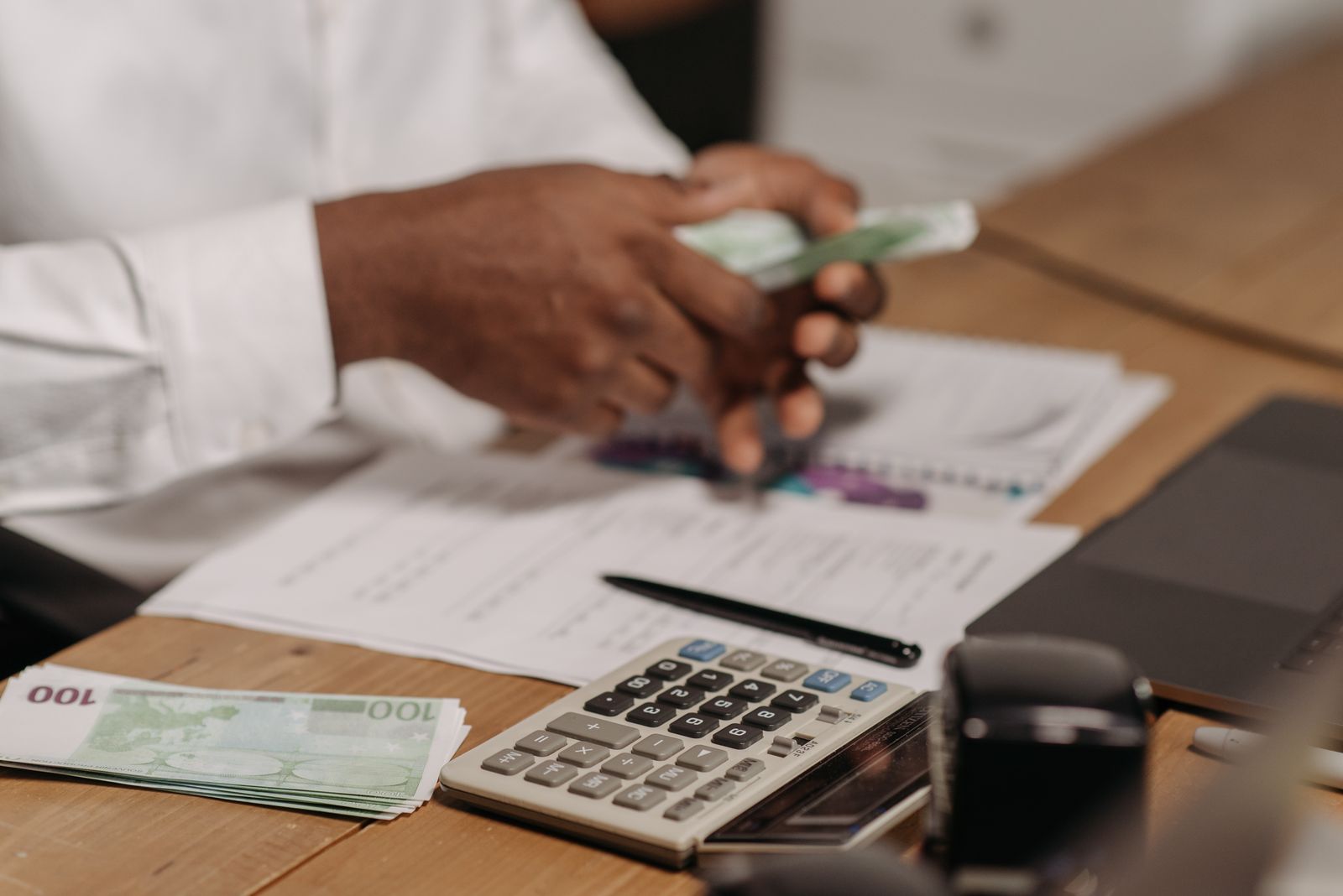 Mâini care numără bancnote pe un birou de lemn, lângă un calculator, acte și un laptop