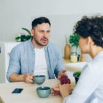 Un bărbat și o femeie stau față în față la masa din bucătărie, cu cești de cafea, într-o discuție serioasă