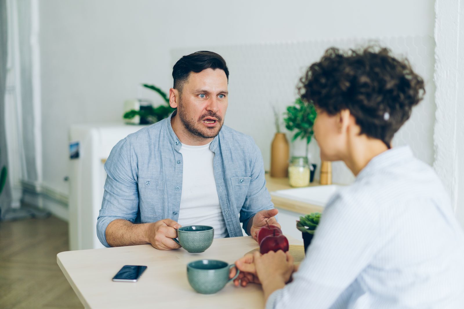 Un bărbat și o femeie stau față în față la masa din bucătărie, cu cești de cafea, într-o discuție serioasă